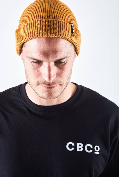 Front view of a man looking downward in a mustard-colored beanie featuring Billy Bones available at CBCO